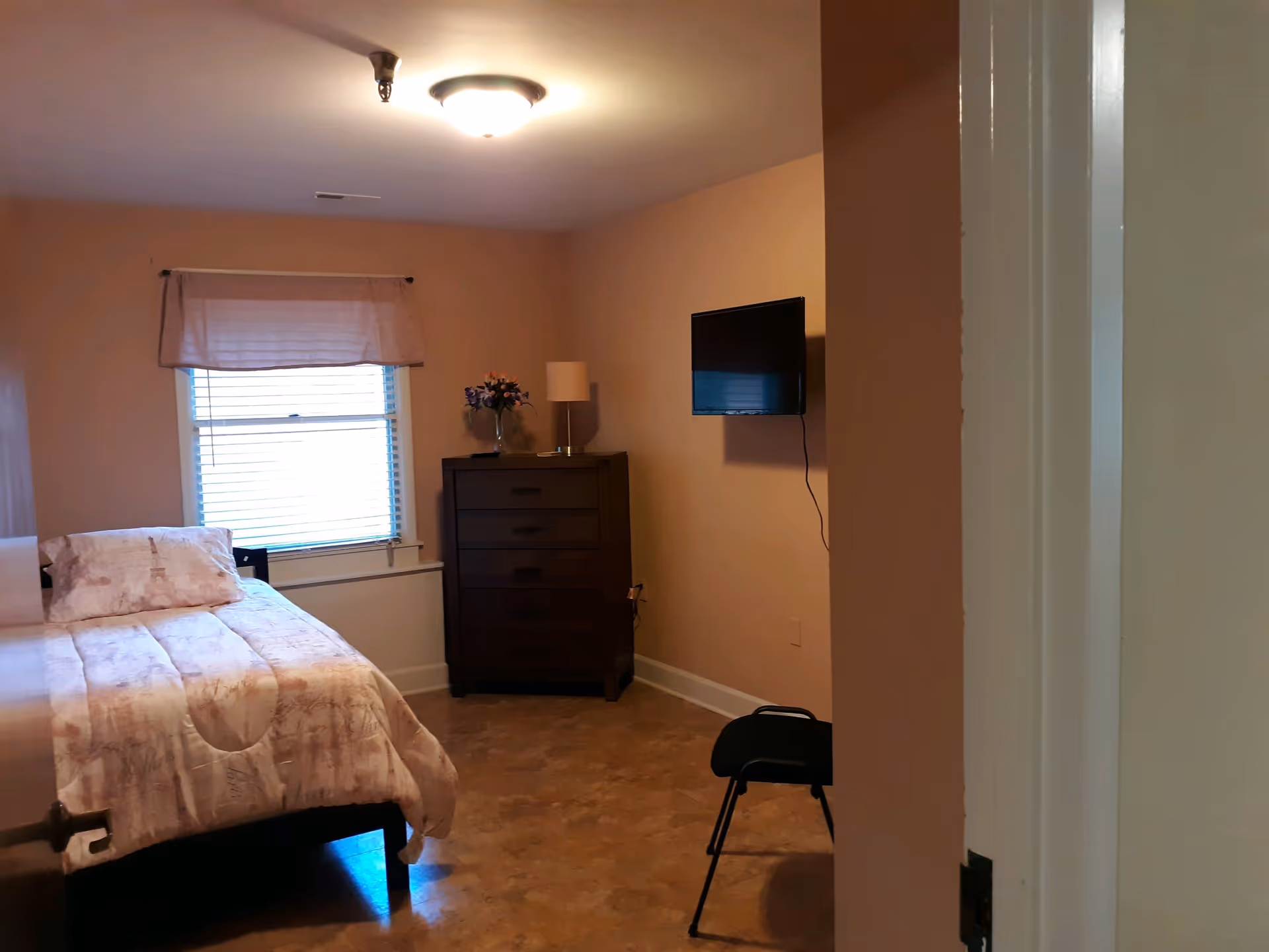A simple bedroom with a bed covered in light-colored bedding, a window with blinds and a valance, a dark wooden dresser with a lamp and a vase of flowers on top, a wall-mounted flat-screen TV, and a black chair near the corner.