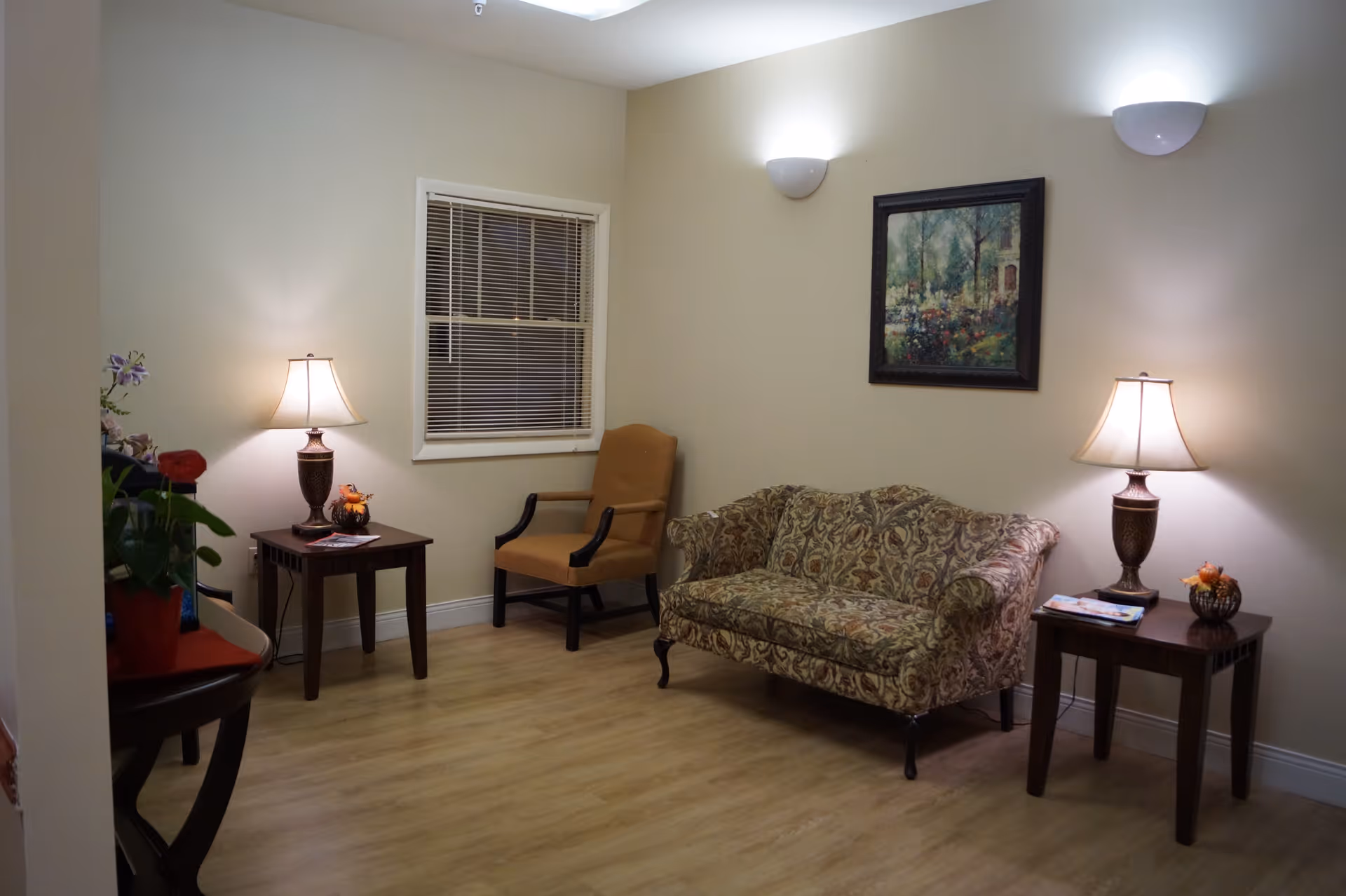 A cozy living room with a patterned sofa, a beige armchair, two wooden side tables each with a lamp, a window with blinds, a framed painting on the wall, and wooden flooring.