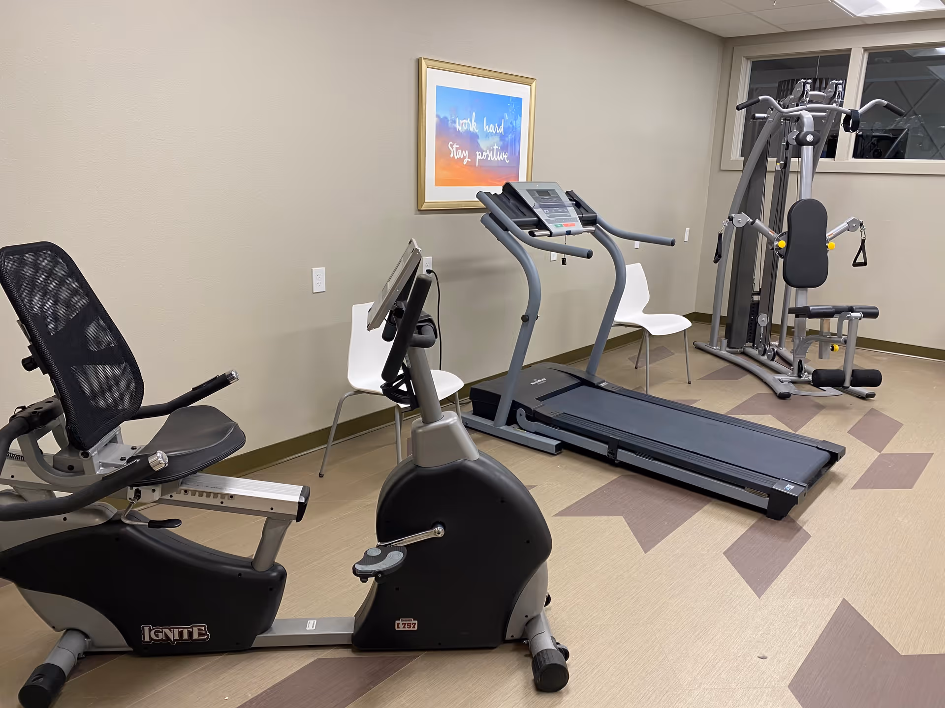 A small fitness room with exercise equipment including a recumbent bike, a treadmill, and a multi-function weight machine. Two white chairs are placed against the wall, and a framed motivational poster that reads 'work hard stay positive' hangs above the treadmill. The room has beige walls and patterned flooring.