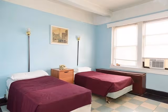 A simple bedroom with two single beds covered in dark red bedspreads, separated by a wooden nightstand with a vintage telephone on it. The walls are painted light blue, and there is a framed picture hanging above the nightstand. Two wall-mounted lamps are positioned above each bed. A large window with blinds and an air conditioning unit is on the right side of the room.