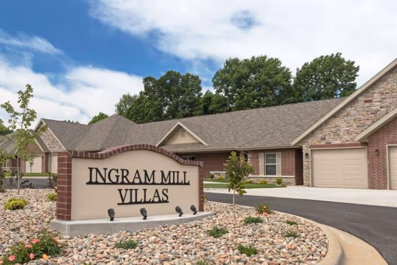 Exterior view of Ingram Mill Villas showing a brick and stone building with attached garages, a curved driveway, and a landscaped area with rocks and small plants surrounding a large sign that reads 'Ingram Mill Villas'. Trees and a partly cloudy sky are visible in the background.