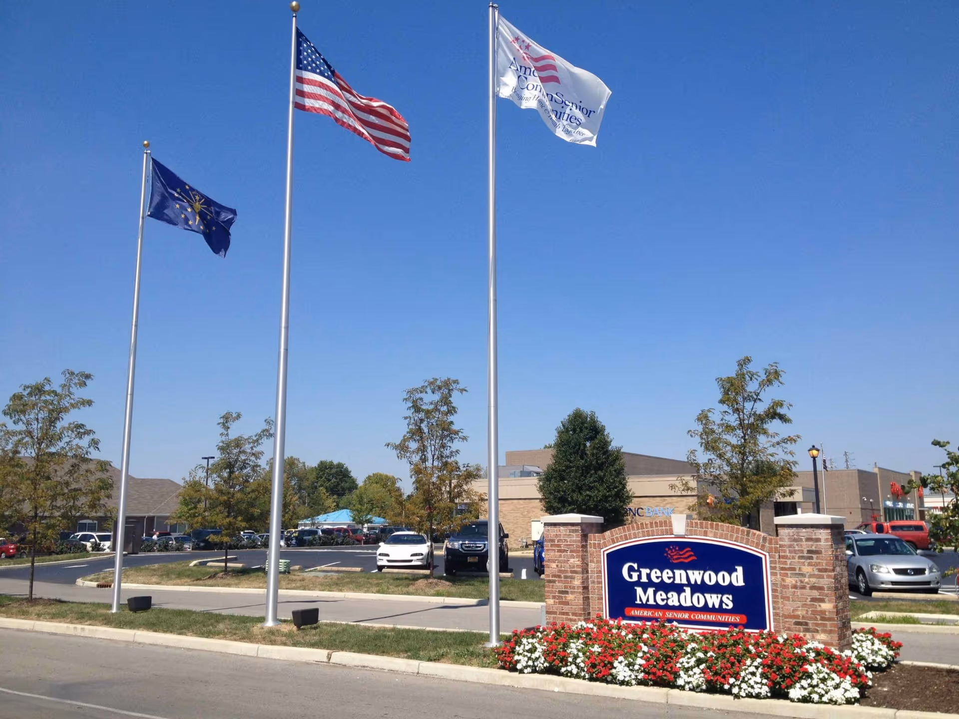 Outdoor view of Greenwood Meadows senior living facility entrance with a brick sign surrounded by red and white flowers. Three flagpoles display the Indiana state flag, the American flag, and a white flag with the American Senior Communities logo. Trees and parked cars are visible in the background under a clear blue sky.