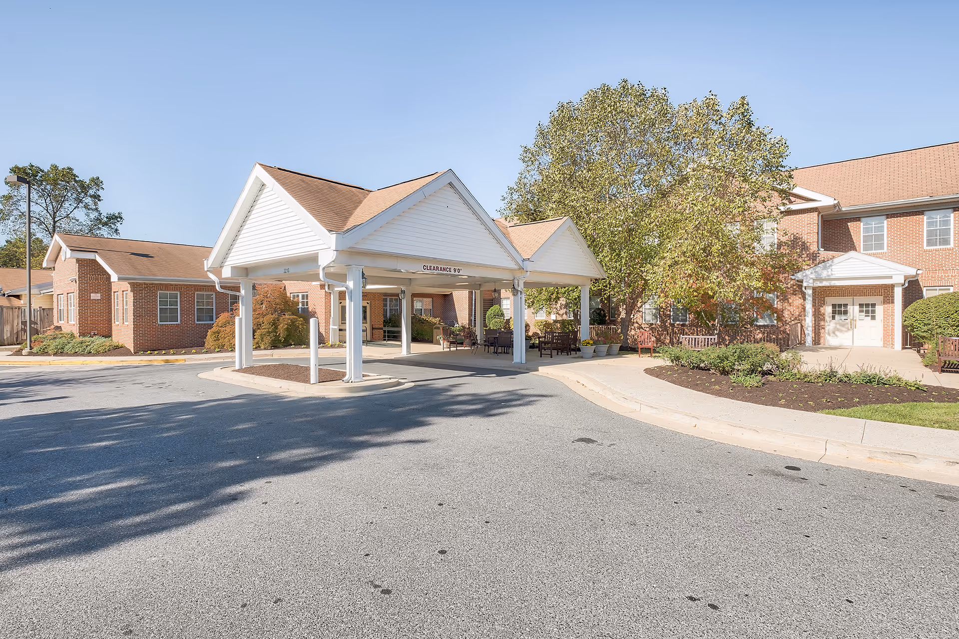 Exterior view of Sterling Care Hillhaven showing a covered entrance with white pillars and a clearance sign. The building is made of red brick with white trim and surrounded by trees and landscaping. There are benches and outdoor seating under the covered area.