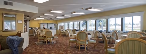 Spacious dining room with multiple round tables and chairs, large windows along one wall, and a serving counter at the far end.