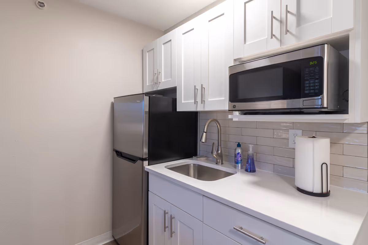 A small modern kitchen area with white cabinets, a stainless steel mini refrigerator, a stainless steel microwave mounted above the counter, a sink with a curved faucet, hand soap and sanitizer bottles, and a paper towel holder on the white countertop.