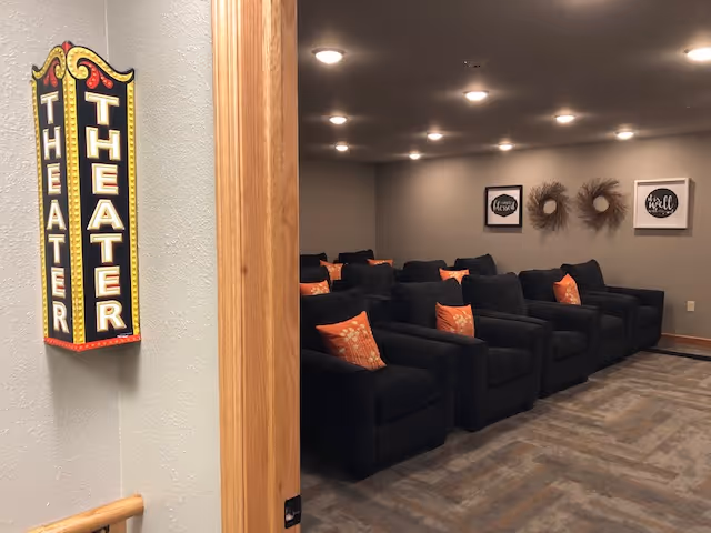 Interior view of a theater room with multiple black cushioned chairs arranged in rows, each chair having an orange pillow with a floral pattern. The walls are gray with two framed decorative art pieces and two wreaths. A vertical sign on the wall near the entrance reads 'THEATER' in bright marquee-style letters.