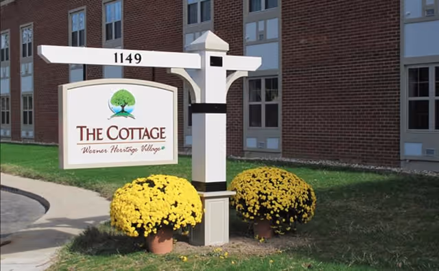 White post sign reading "The Cottage" and address 1149 in front of a brick building with two yellow potted mums.