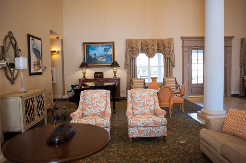 A spacious living room area with two floral patterned armchairs facing a round wooden table with a decorative sculpture. Behind the chairs, there is a grand piano with two lamps and a framed painting on the wall. To the right, there is a window with draped curtains and a small dining table with four chairs. The room has beige walls, a patterned carpet, and a large white column near the entrance door.