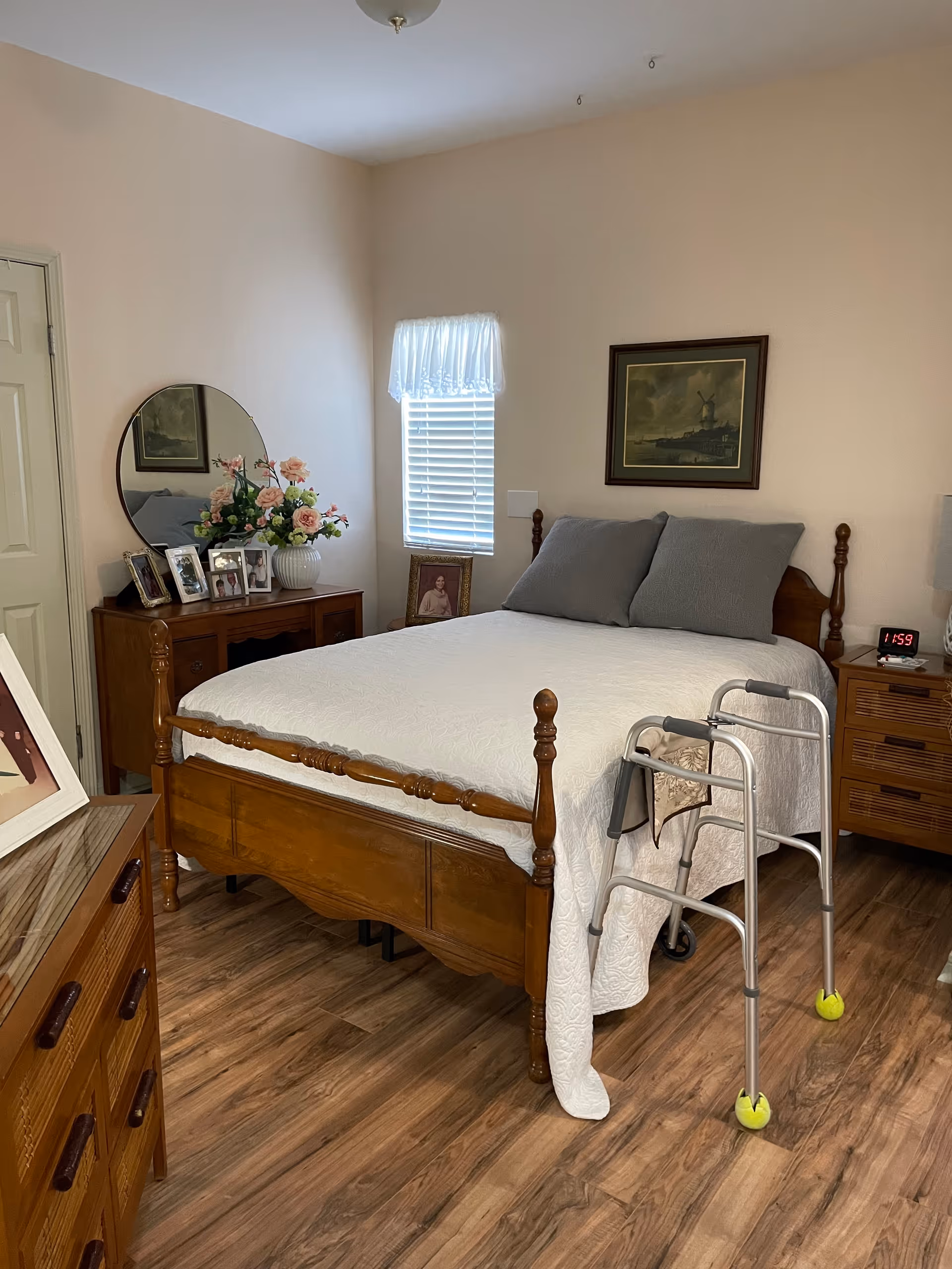 A cozy bedroom with a wooden bed frame covered with a white quilt and two gray pillows. A walker with tennis balls on the back legs is positioned at the foot of the bed. There is a wooden dresser with a round mirror, a vase of flowers, and framed photos on top. A small window with white blinds and a valance is on the wall behind the bed, along with a framed picture. The floor is wood, and a digital clock on a nightstand shows the time 1:59.