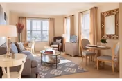Bright, neutrally decorated living room with a sofa, armchairs, coffee table, small dining table, and large windows with beige curtains.