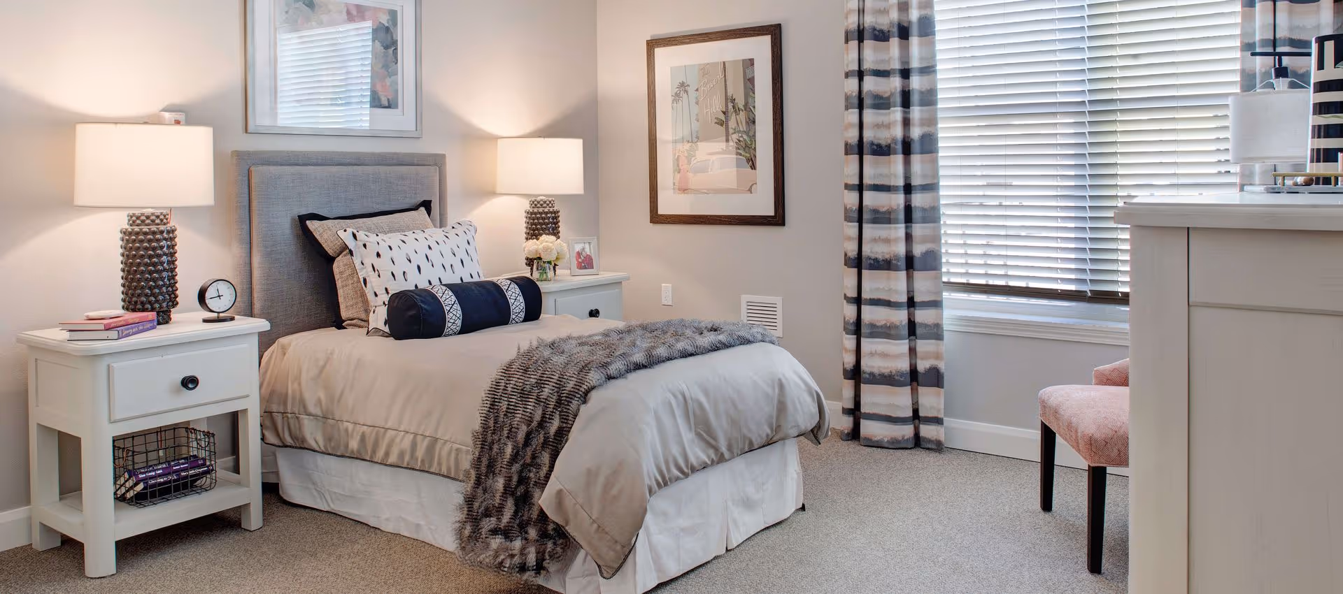A neatly arranged bedroom with a single bed featuring beige bedding, decorative pillows, and a gray throw blanket. There are two white nightstands on either side of the bed, each with a lamp and decorative items. A framed picture hangs on the wall above the bed, and a window with blinds and striped curtains is visible on the right side. A pink upholstered chair is partially visible near a white dresser.