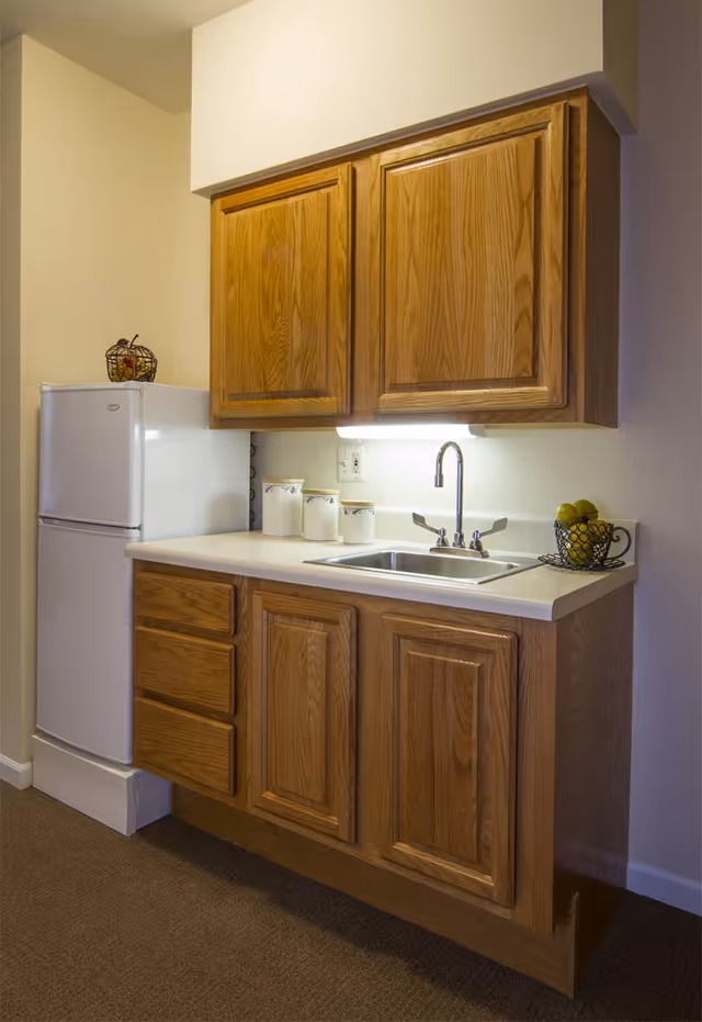Small kitchen area with wooden cabinets, a white countertop, a stainless steel sink with a faucet, a white refrigerator, and decorative containers and a fruit basket on the counter.