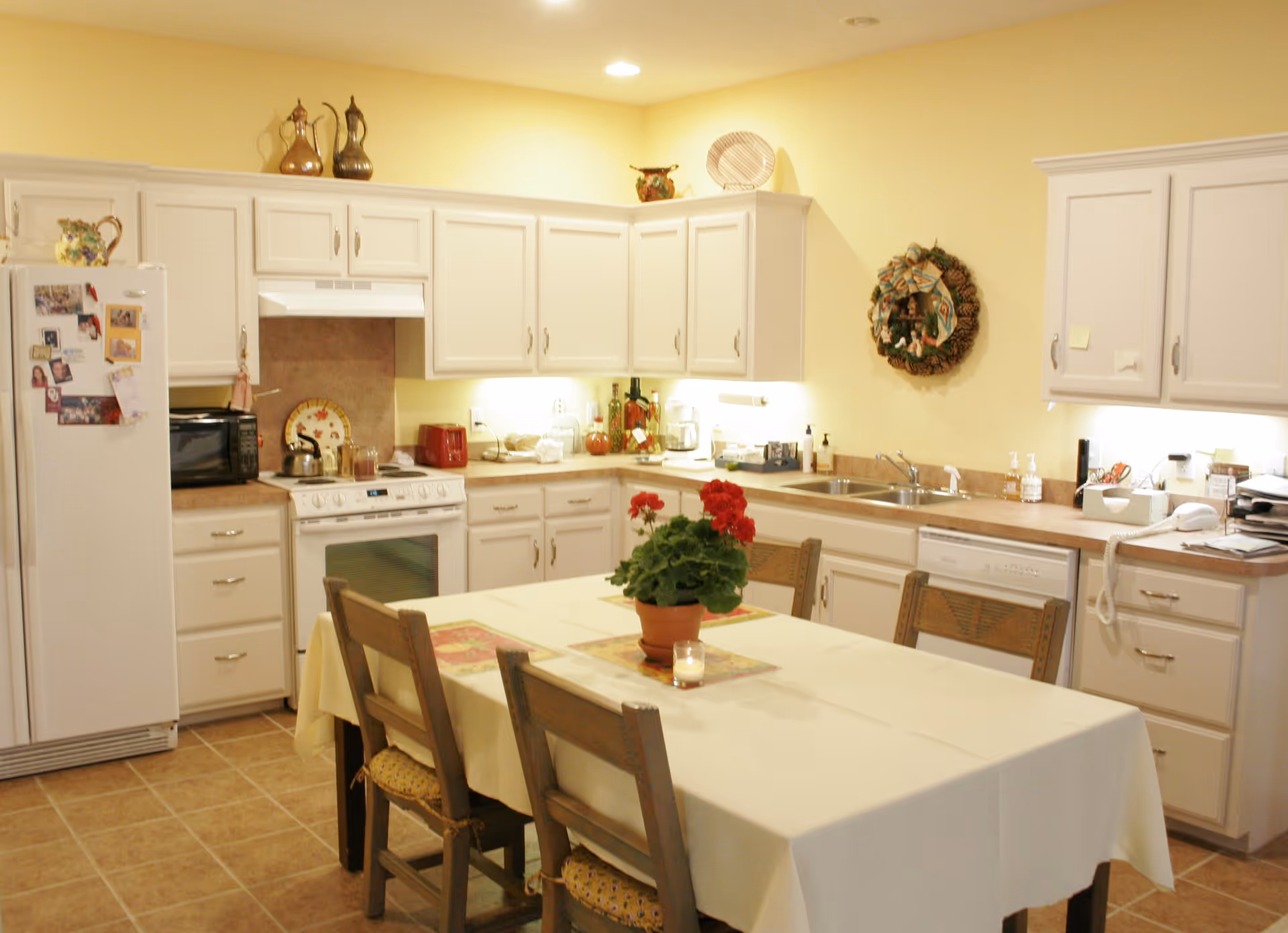 A bright kitchen with white cabinets, a white refrigerator covered with photos and magnets, a white stove, a microwave, and a dishwasher. A table with a white tablecloth and four wooden chairs is in the center, decorated with a potted red flower and a small candle. The walls are painted yellow, and there is a decorative wreath hanging above the sink area.