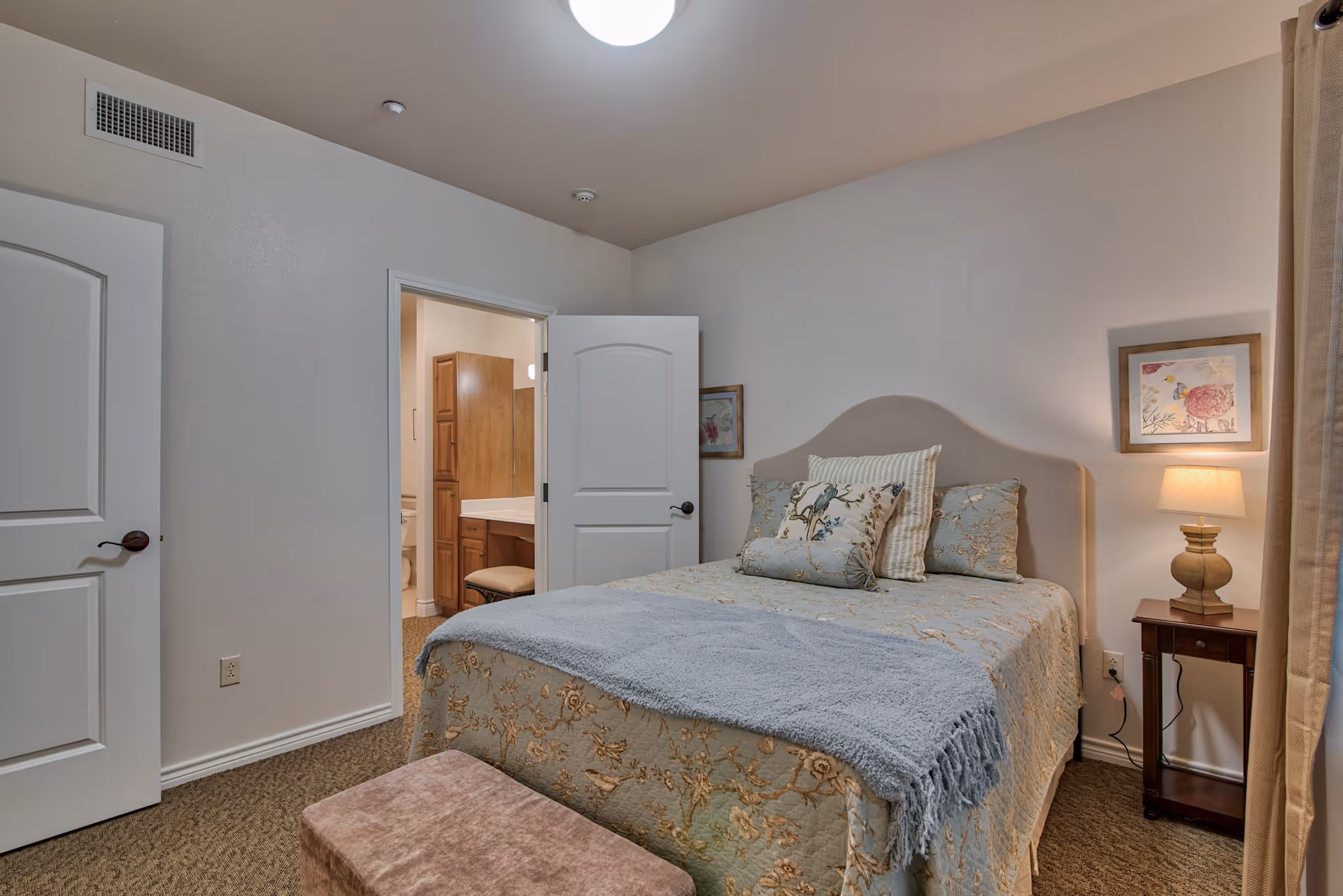 A cozy bedroom with a bed covered in floral bedding and multiple pillows. There is a small wooden nightstand with a lamp and framed artwork on the wall. The room has beige walls, carpeted floor, and an open door leading to a bathroom with wooden cabinets and a vanity.