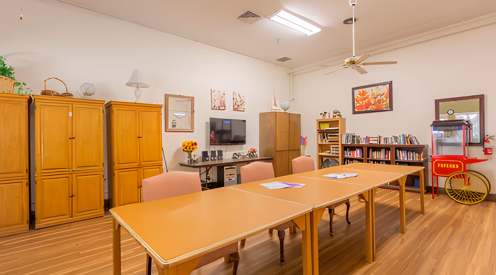 A well-lit room with wooden flooring featuring a long table with several chairs around it. The walls have wooden cabinets, a TV mounted on the wall, bookshelves filled with books, framed artwork, and a red popcorn machine. A ceiling fan and fluorescent light fixture are visible on the ceiling.