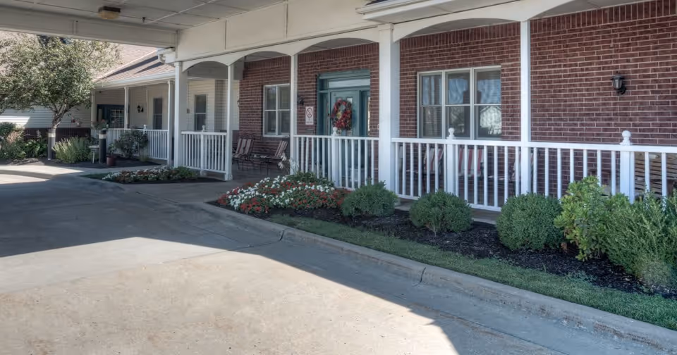 Covered porch area of a senior living facility with white railings, brick walls, and a green door decorated with a wreath. There are chairs on the porch and flower beds with shrubs and flowers along the edge. A tree and additional building structures are visible in the background.