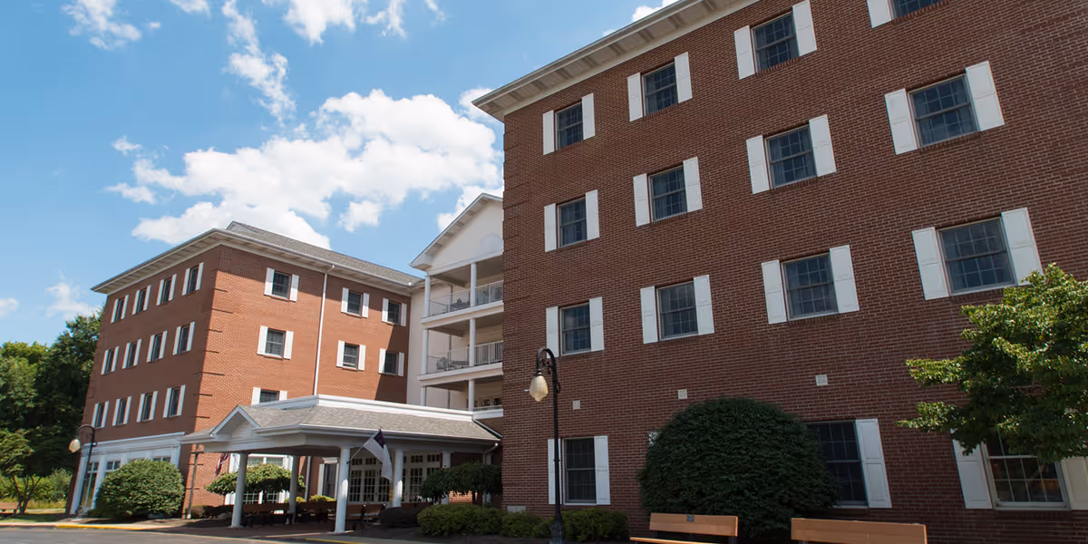 Multi-story red-brick residential building with a covered entrance, benches, and landscaping under a blue sky.
