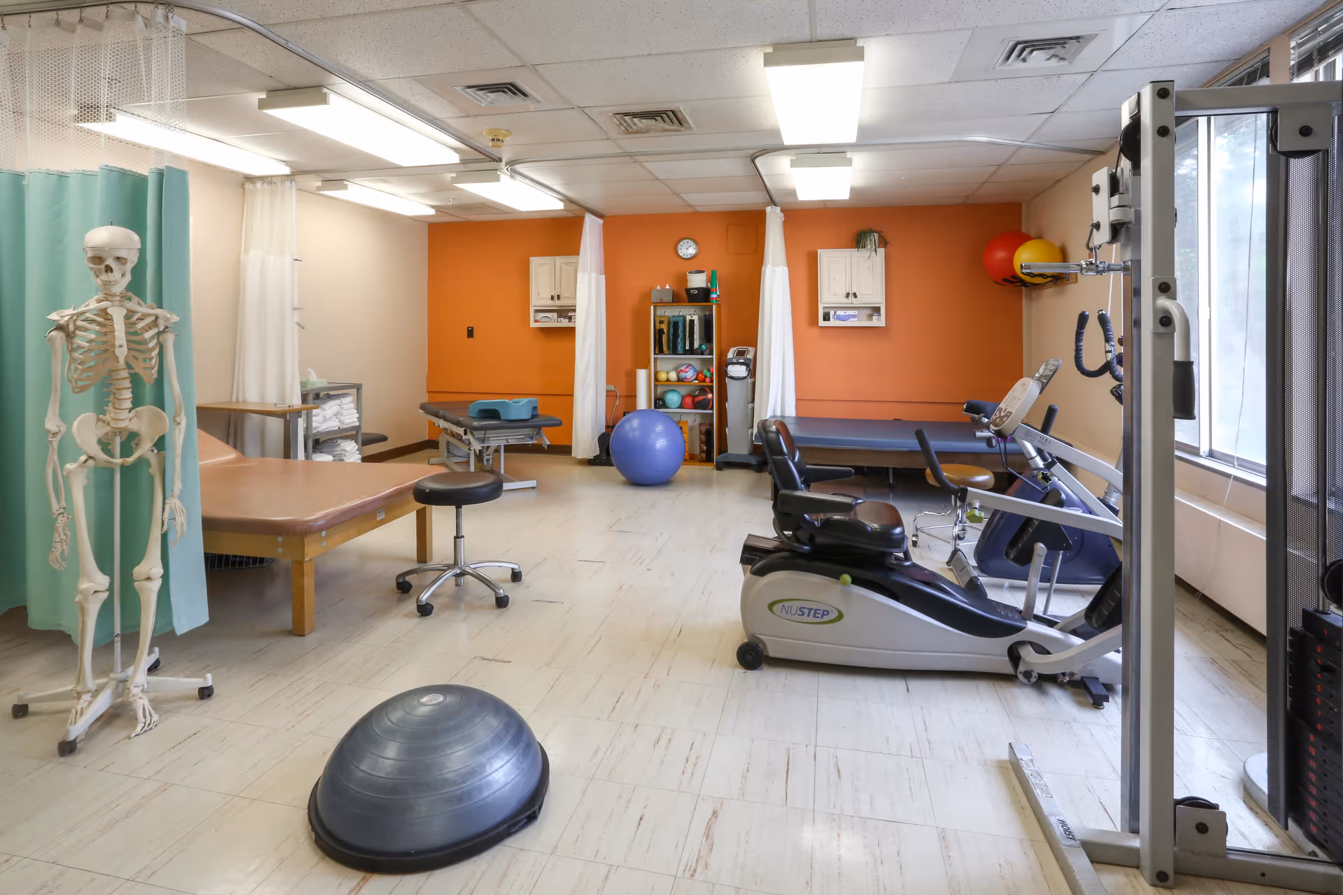 A physical therapy or rehabilitation room with exercise equipment including a recumbent bike, therapy tables, exercise balls, and a skeleton model. The room has orange accent walls, white ceiling tiles with fluorescent lighting, and large windows letting in natural light.