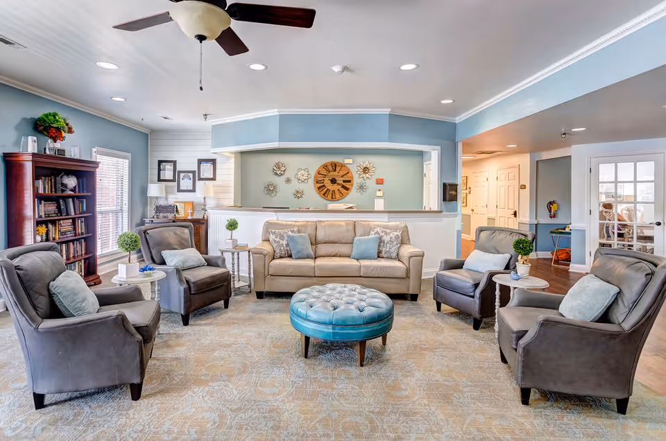 A cozy living room area in a senior living facility with a beige sofa, four gray armchairs with light blue cushions, a round blue tufted ottoman in the center, a wooden bookshelf filled with books, and decorative wall art including a large clock and floral metal pieces. The walls are painted light blue with white trim, and the room is well-lit with natural light from windows and ceiling lights.