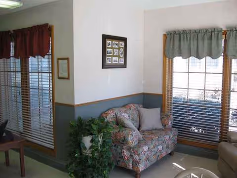 Senior living facility living room with a floral sofa, large windows, blinds, plant, and wall photos.