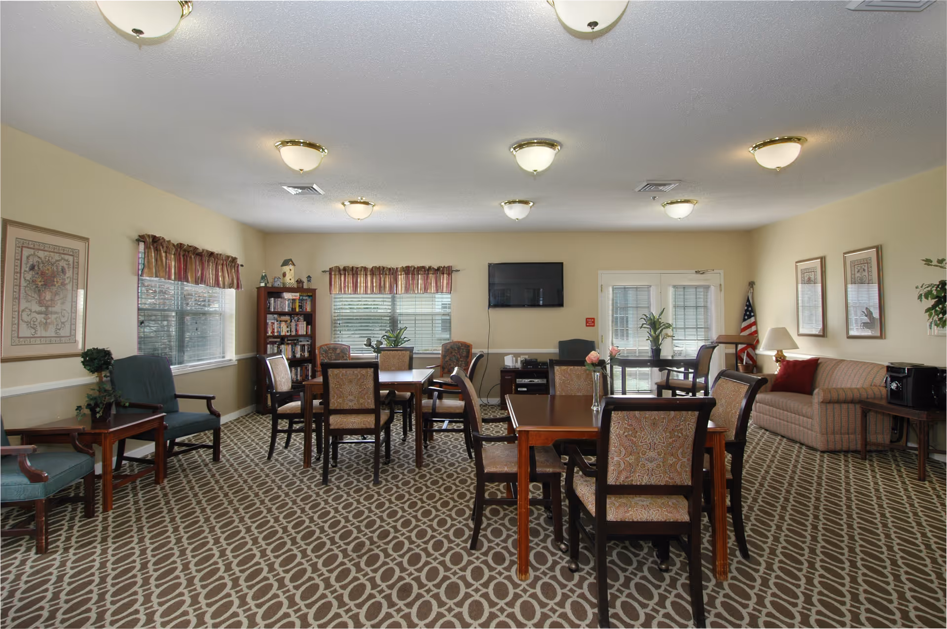 A well-lit common area with patterned carpet, several wooden tables and chairs, a beige couch, framed artwork on the walls, windows with valances, a bookshelf, and a wall-mounted TV. There is also an American flag in the corner and a door with glass panels.