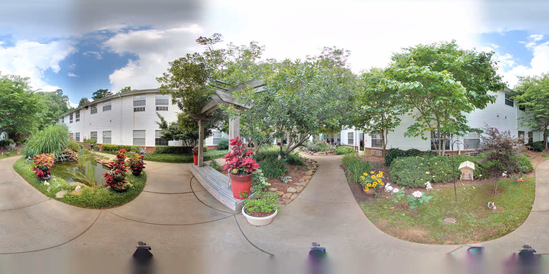 Outdoor garden area at Oaks at Tucker featuring a paved walkway surrounded by lush greenery, colorful flowers, small trees, and a small pond. The white exterior of the building is visible in the background under a partly cloudy sky.