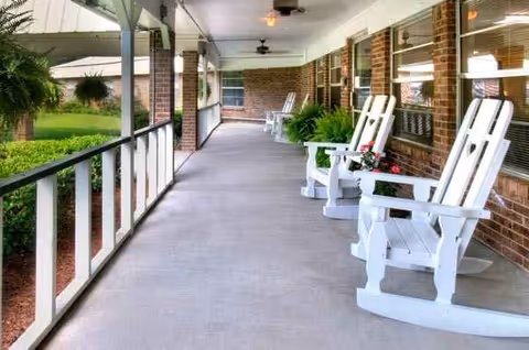 Covered outdoor porch with white rocking chairs along brick building, plants nearby.