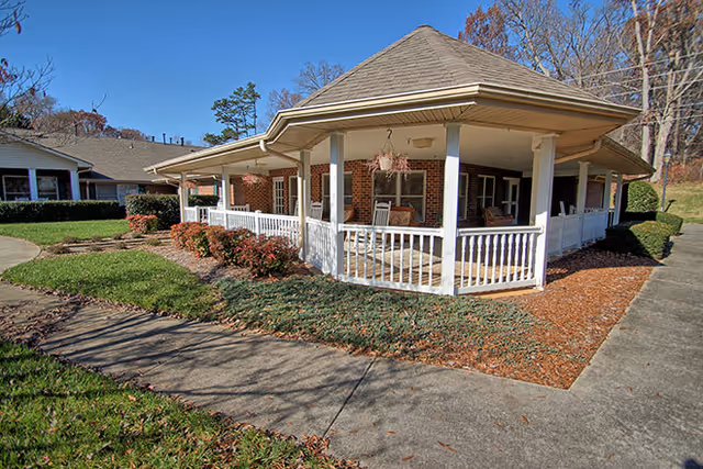 Brick senior living building with a covered wraparound porch, white railing, rocking chairs, landscaping and a concrete walkway.