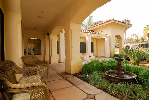Covered outdoor walkway with wicker chairs on the left and a small garden with a water fountain on the right, adjacent to a building with beige walls and a red-tiled roof.