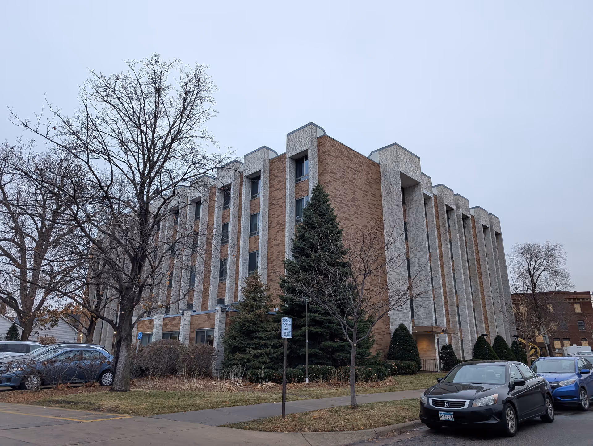 Exterior view of a multi-story brick and concrete building with several windows, surrounded by leafless trees and parked cars on a cloudy day.