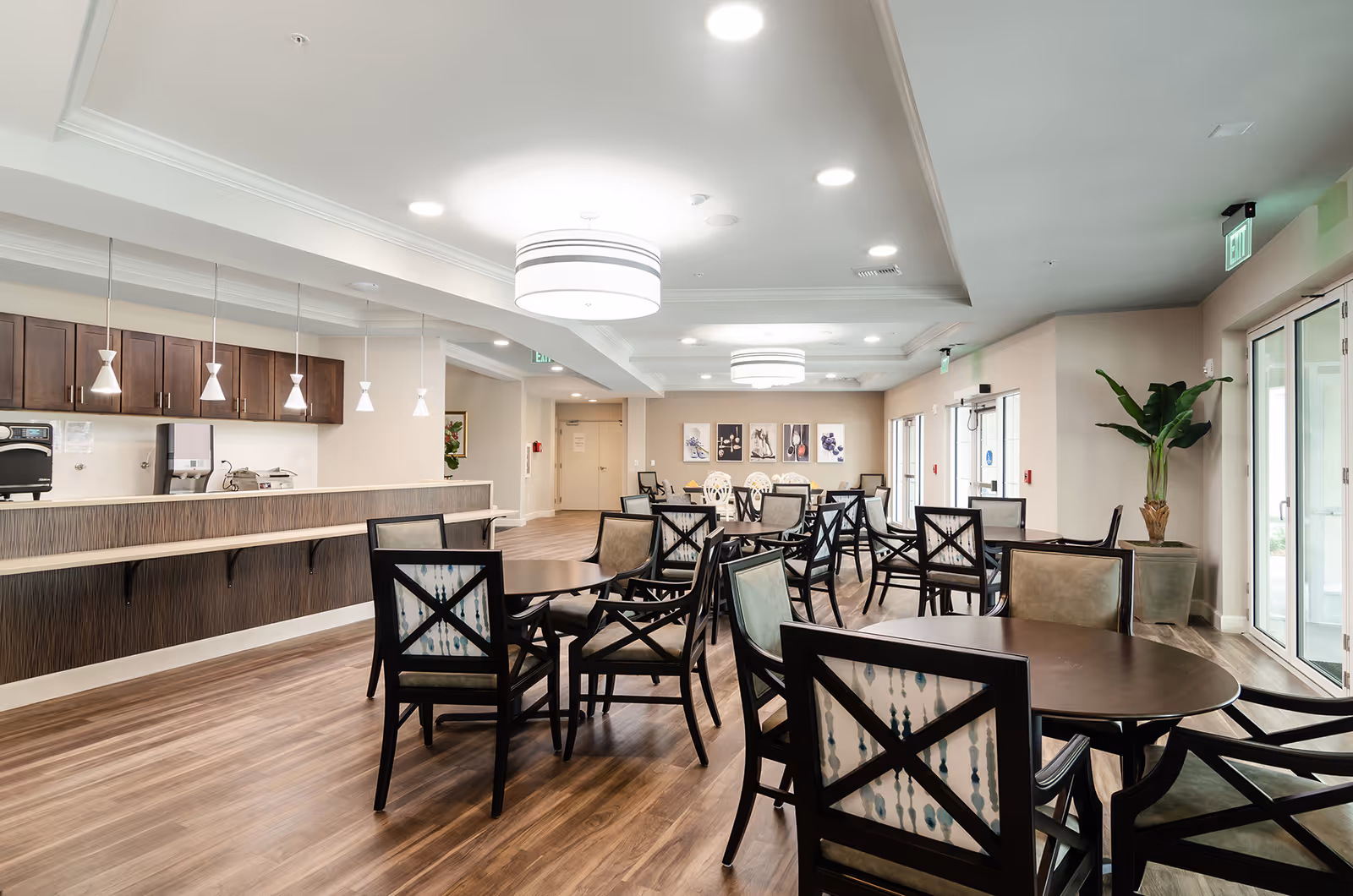 A spacious dining area with multiple round tables and chairs arranged neatly. The room features wooden flooring, modern pendant and ceiling lights, a counter with cabinets and a beverage dispenser, and large windows allowing natural light. There is a potted plant near the entrance and framed artwork on the far wall.