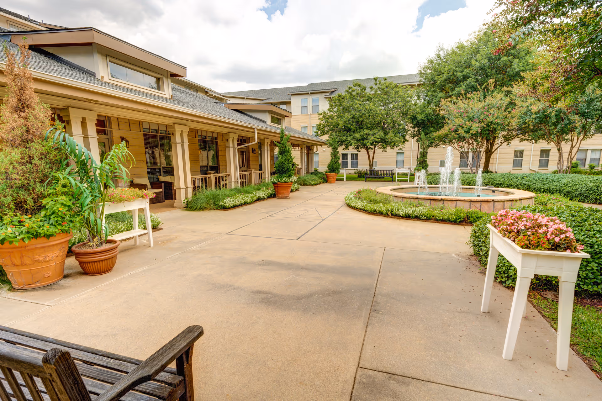 Outdoor courtyard area at Town Village Crossing featuring a paved walkway, potted plants, a water fountain with multiple streams, benches, and a building with a covered porch in the background under a partly cloudy sky.