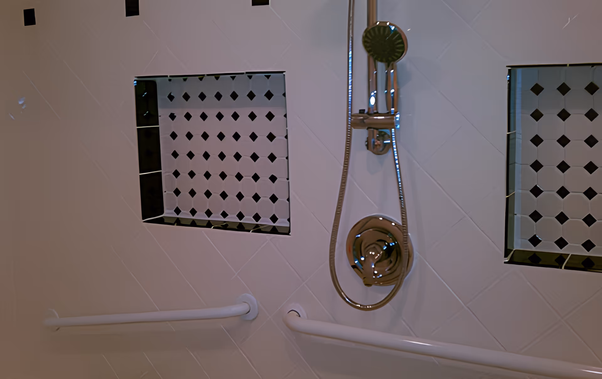 Close-up view of a shower wall with white tiles, featuring two recessed shelves with black and white patterned tiles, a chrome showerhead with an adjustable hose, and two white grab bars installed horizontally on the wall.
