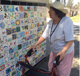 An elderly woman wearing a straw hat, glasses, a white shirt, and pink pants is standing outdoors using a walker. She is looking at a colorful tiled mural on a wall, which features various artistic designs and drawings.