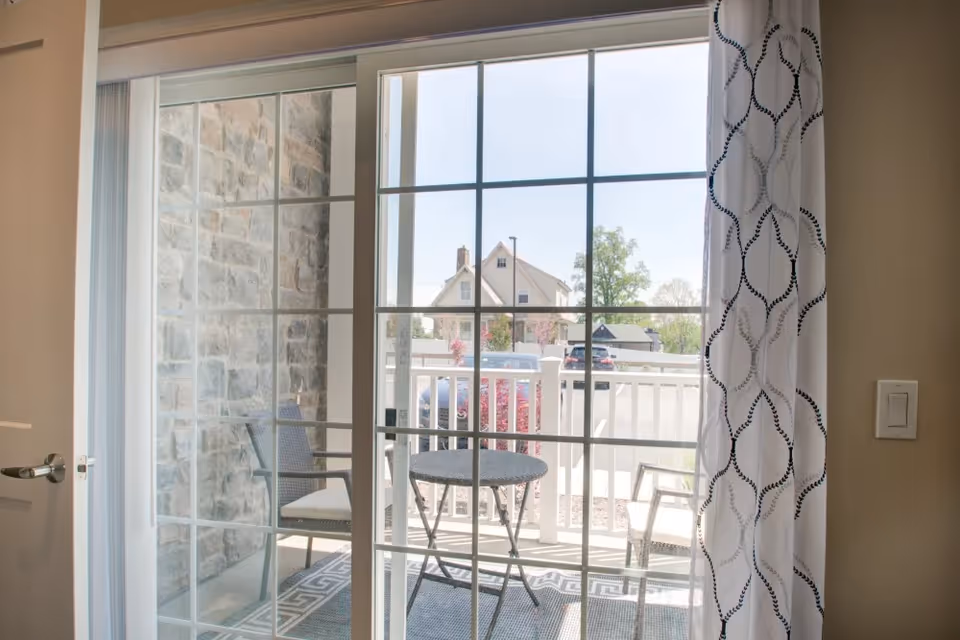 View through a glass sliding door with white grid frames, showing a small outdoor patio area with a round table and two chairs. The patio has a patterned rug and is enclosed by a white railing. Outside, there is a stone wall on the left and a parking area with cars and a house in the background.