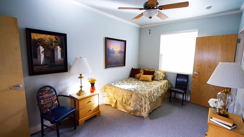 A cozy bedroom with a single bed covered in a gold patterned bedspread and several pillows. The room has light blue walls, two wooden doors, a ceiling fan with light, two framed paintings on the walls, a wooden nightstand with a lamp and a vase of flowers, a wooden dresser with a lamp and books, and two chairs, one with a patterned cushion and one plain black.