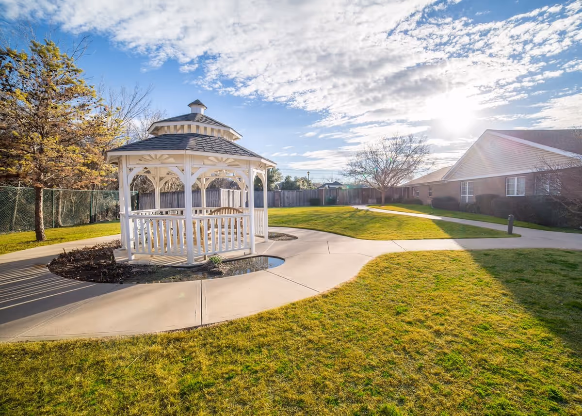 Outdoor area of a senior living facility with a white wooden gazebo surrounded by a concrete pathway, green grass, trees, and a building in the background under a partly cloudy sky with the sun shining.