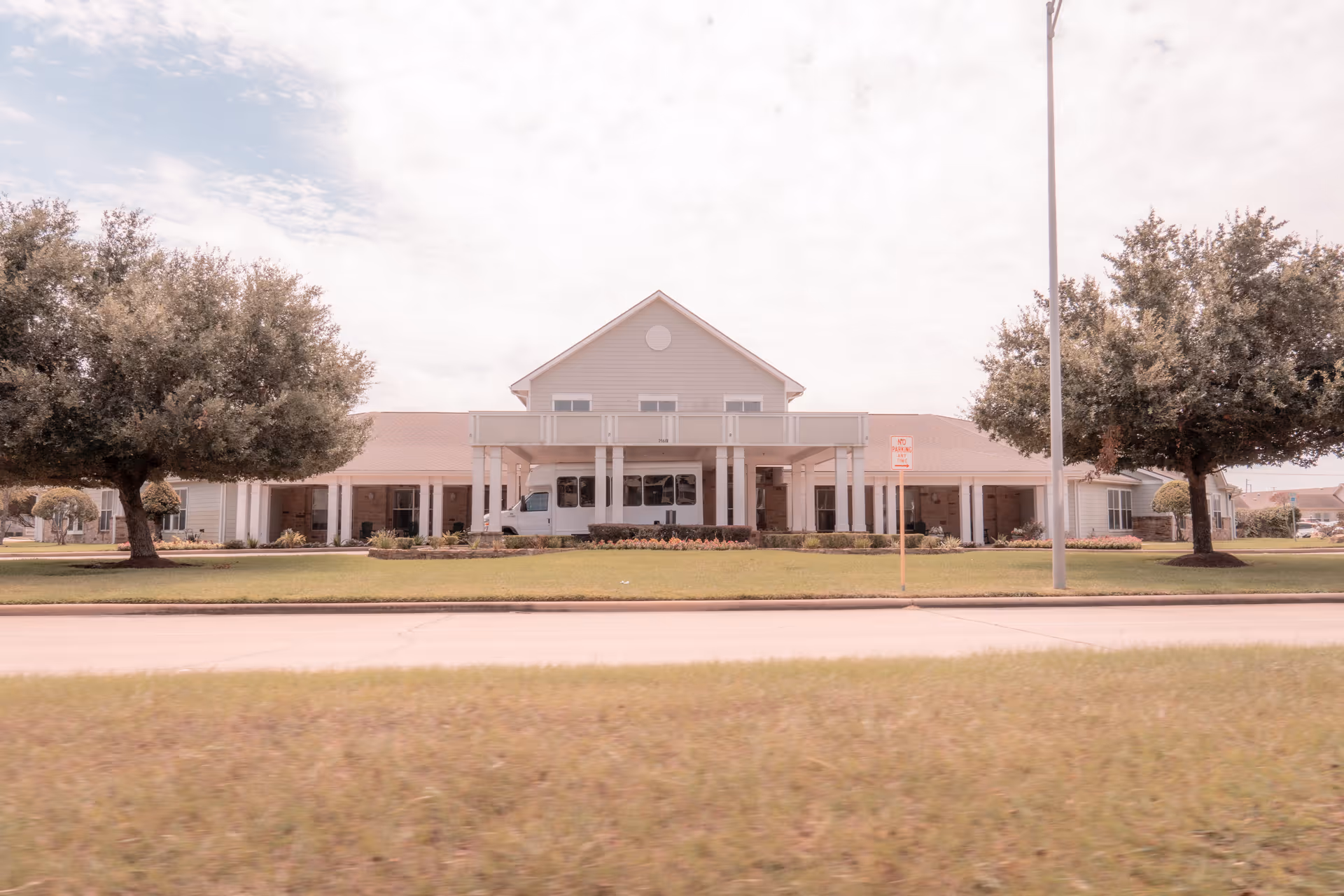 Front exterior of a single-story assisted living building with a covered entrance, driveway, lawn, and trees.