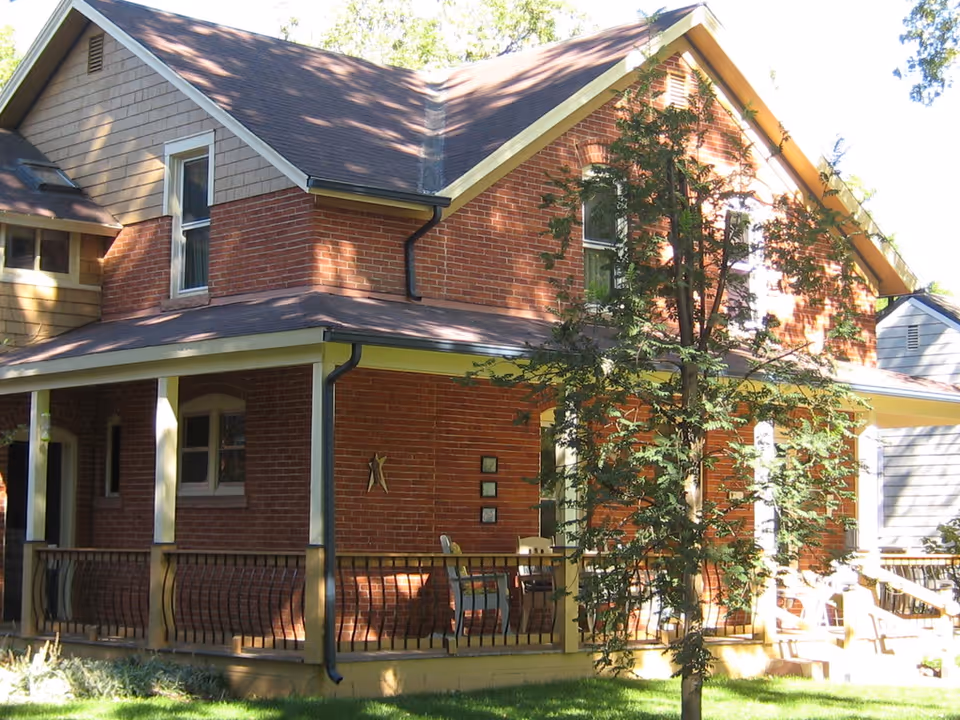 Exterior view of a two-story brick house with a covered porch, railing, and outdoor chairs. A tree is in the foreground with sunlight casting shadows on the building.