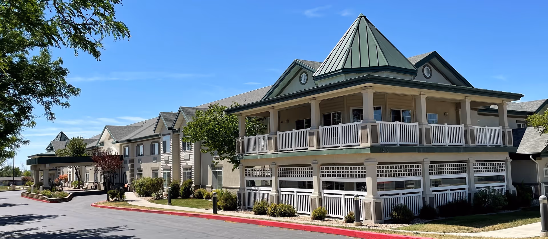 Exterior view of a large, two-story assisted living facility building with a green metal roof and white railings on the upper balcony. The building is surrounded by well-maintained landscaping including bushes and trees, with a paved driveway and clear blue sky in the background.
