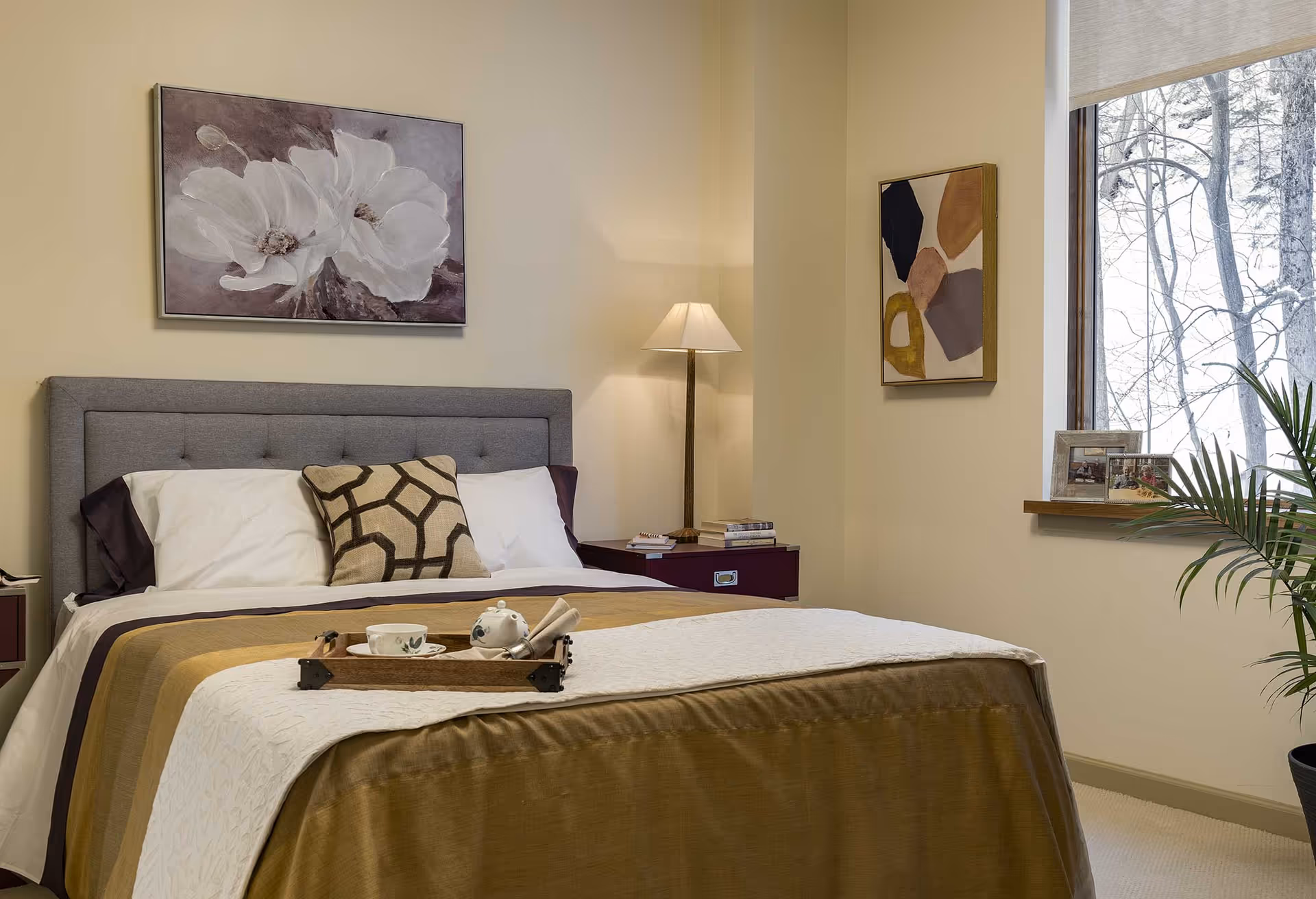 A neatly made bed in a softly lit bedroom with framed artwork, bedside tables, a lamp, and a window showing trees outside.