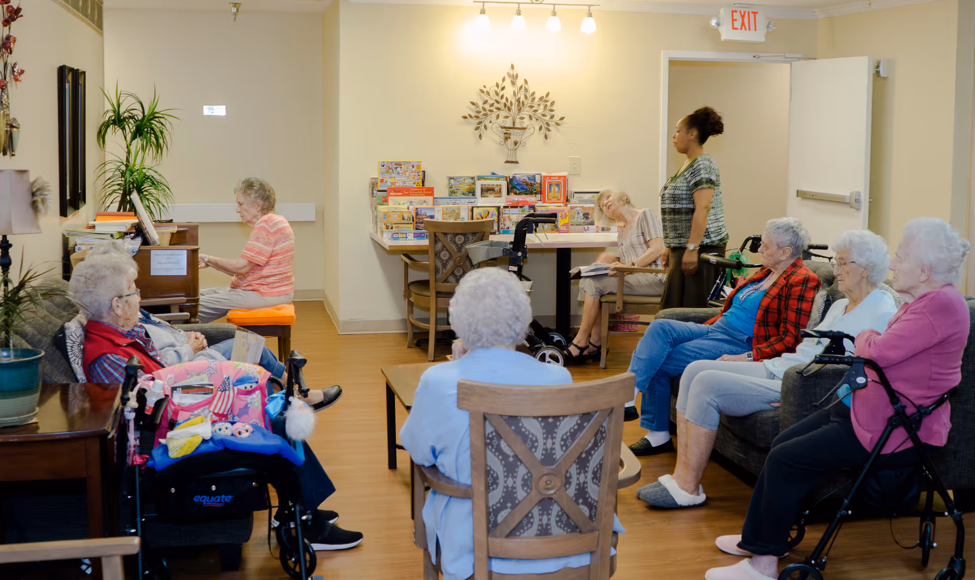 A group of elderly women sitting in a common room at a retirement center. One woman is playing the piano while others are seated on chairs and sofas, some with walkers nearby. A staff member stands near the back of the room. The room has wooden floors, a table with puzzles, and wall decorations.