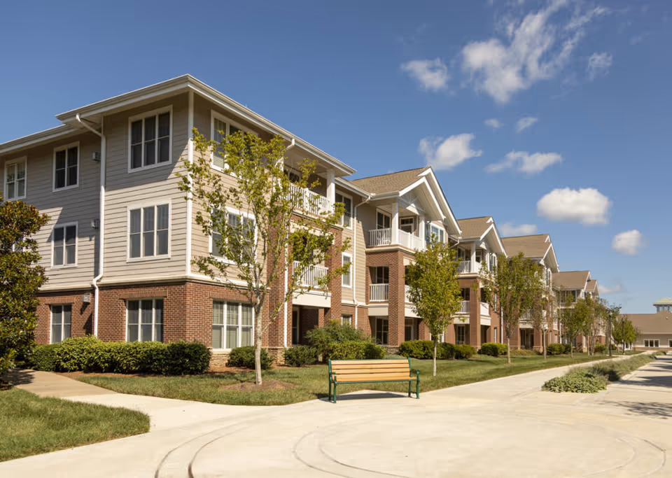 Exterior view of a multi-story senior living facility building with beige siding and red brick accents, surrounded by small trees and shrubs under a blue sky with scattered clouds. A wooden bench is placed on a paved walkway in front of the building.