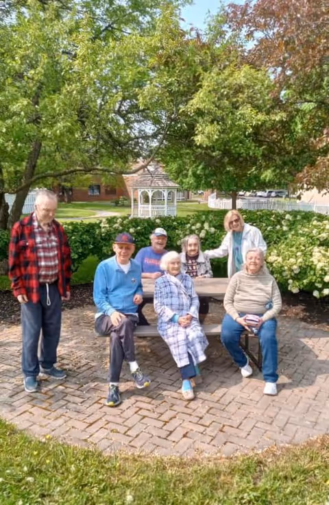 A group of six elderly people and one caregiver outdoors in a garden area with green trees, bushes, and a white gazebo in the background. Four elderly individuals are seated on a wooden bench and a chair on a brick-paved area, while two elderly men stand nearby. The caregiver stands behind the bench, smiling and interacting with the group.