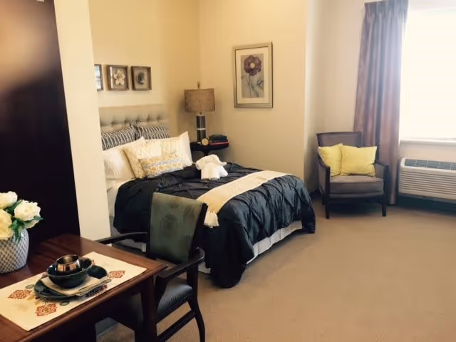 A cozy bedroom in a senior living facility featuring a neatly made bed with black and white bedding, a bedside table with a lamp, a framed flower picture on the wall, a comfortable armchair with yellow pillows near a window with brown curtains, and a small dining table with two chairs and a flower vase.