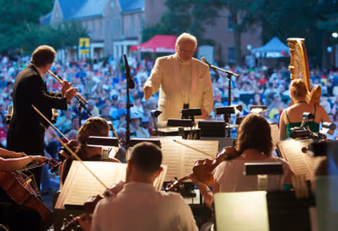 Conductor leading an outdoor orchestra performance with musicians and a large seated audience in the background.