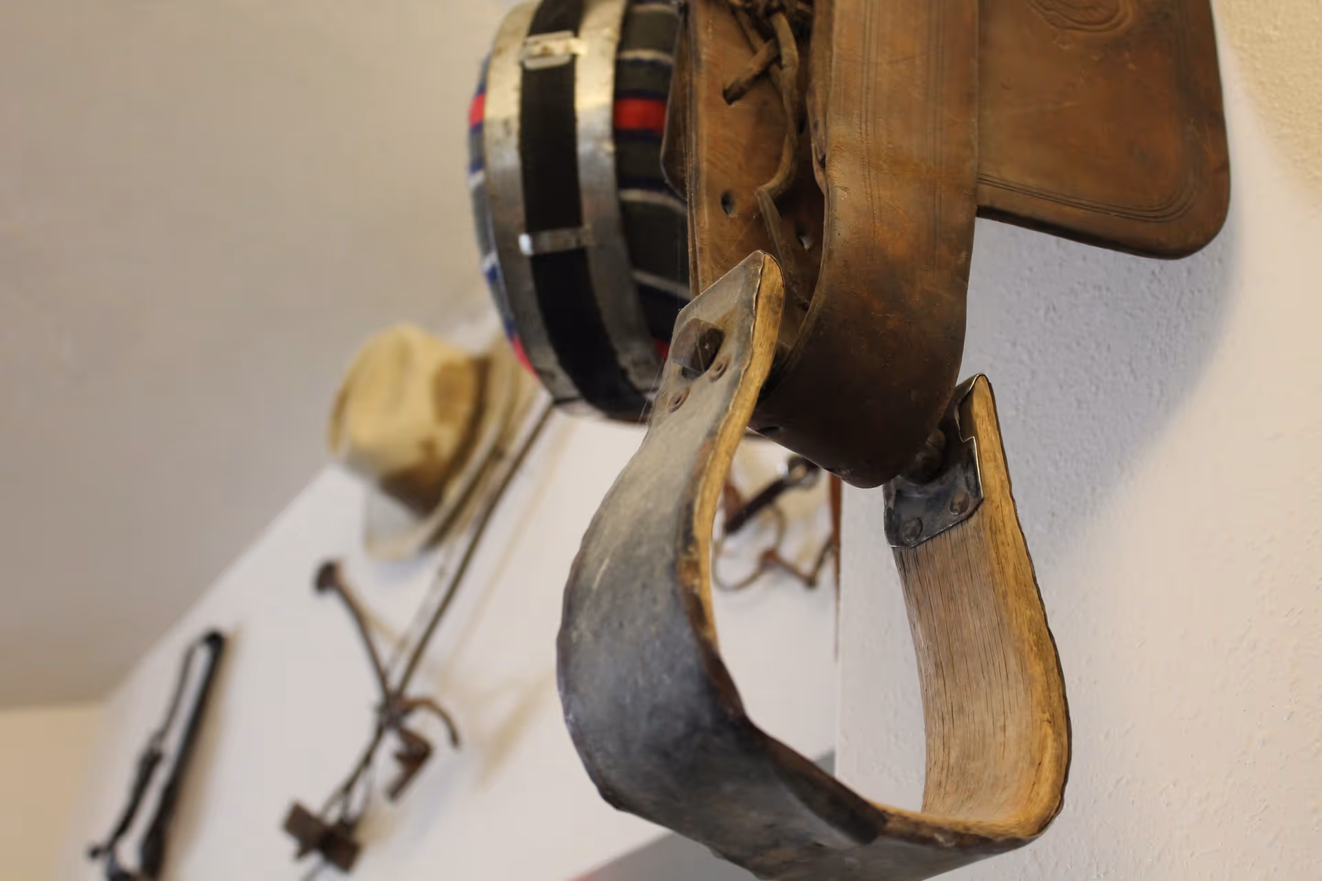 Close-up view of vintage horse tack and a hat hanging on a wall, including a leather saddle and a wooden stirrup, with other equestrian equipment in the background.
