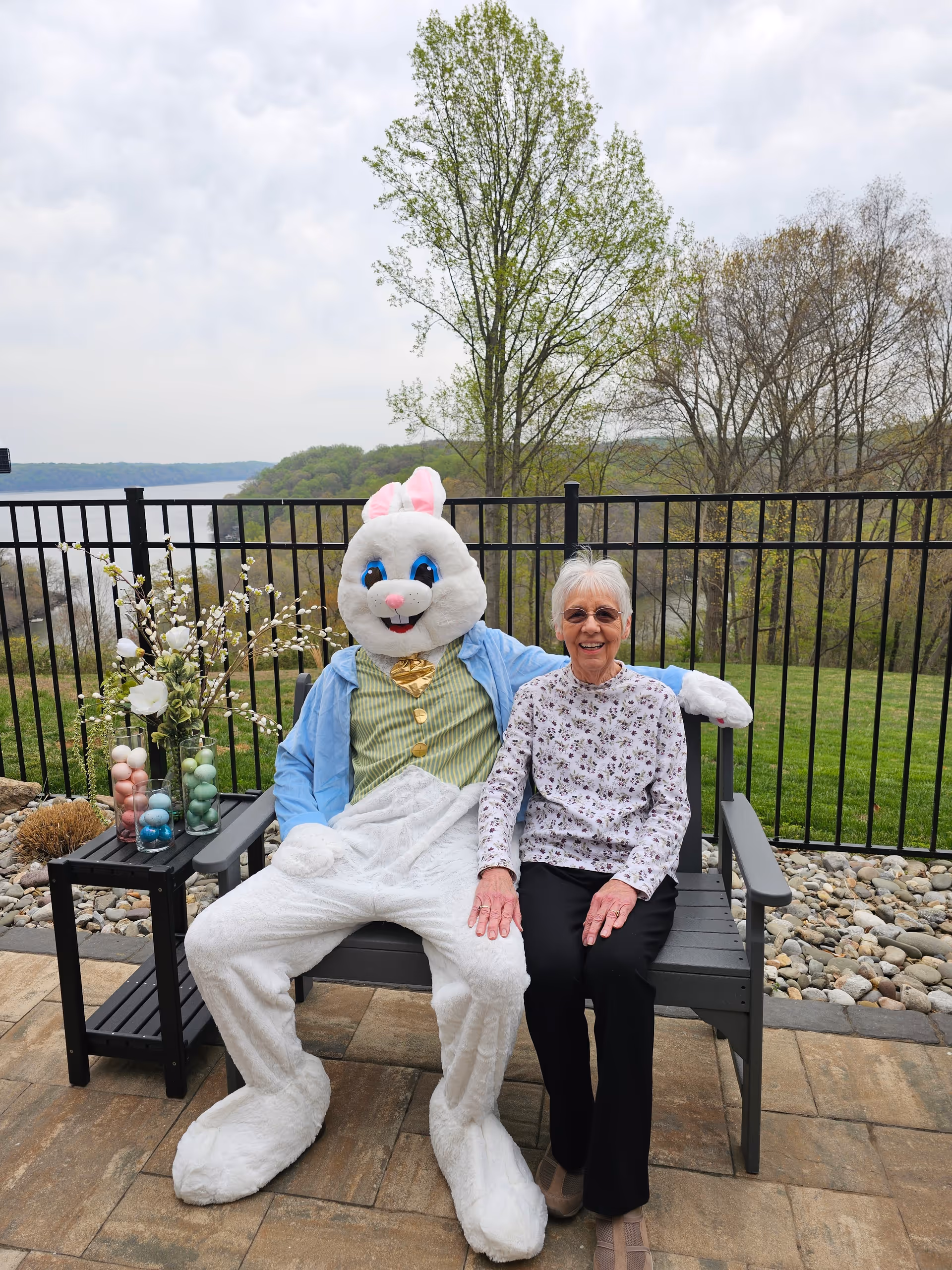 An elderly woman sitting on a bench outdoors next to a person dressed in a large white Easter Bunny costume. They are smiling and holding hands. Behind them is a black metal fence, green grass, trees, and a body of water in the distance. A small table next to the bench holds vases with decorative branches and pastel-colored Easter eggs.