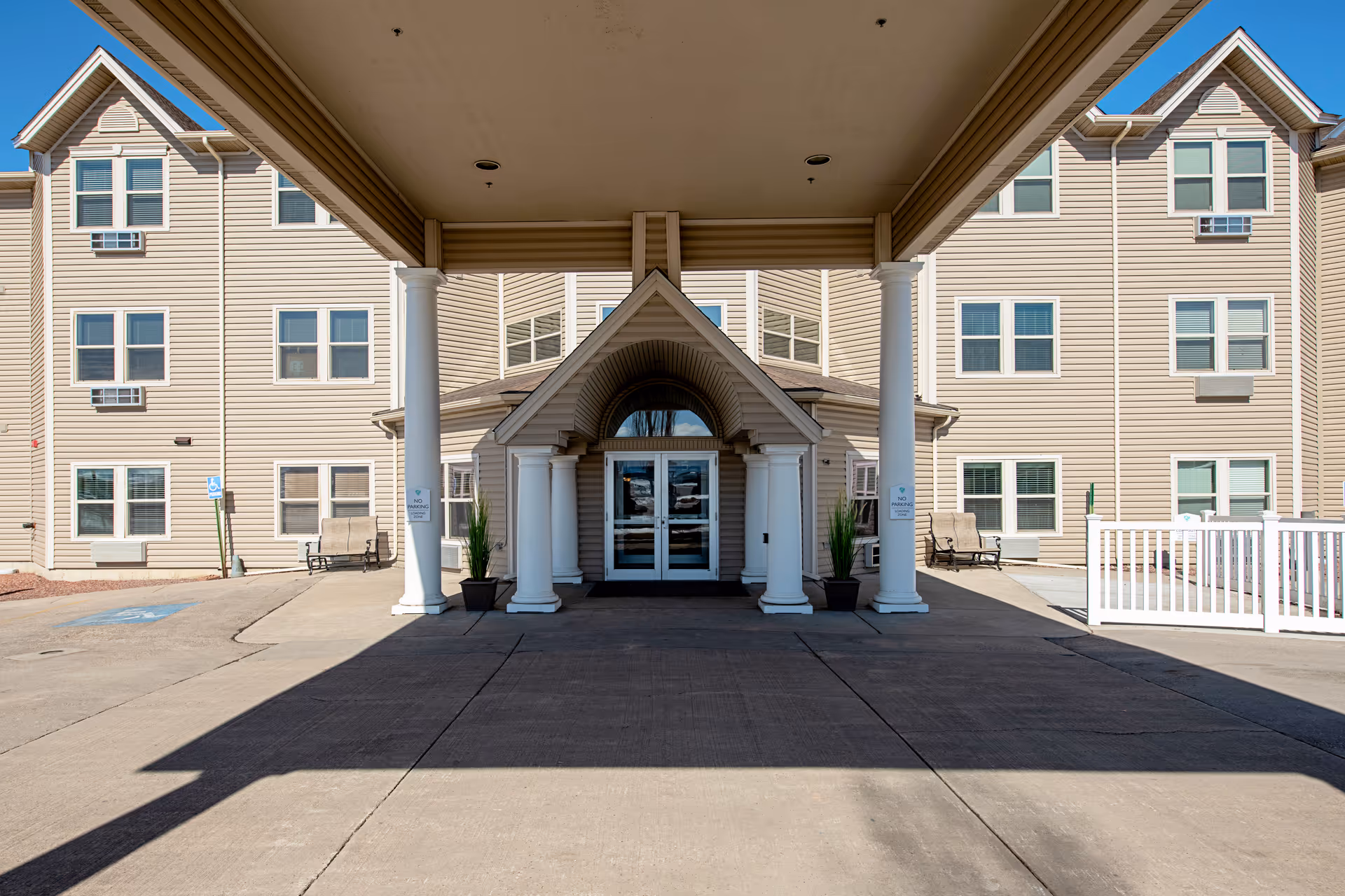 Entrance of a senior living facility with a covered driveway supported by white columns. The building has beige siding, multiple windows, and benches on either side of the entrance. There are potted plants near the doorway and a clear blue sky above.