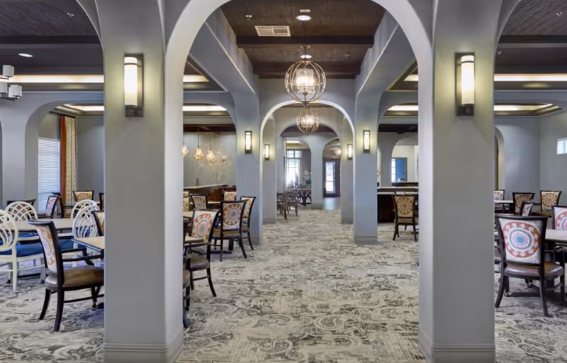 Spacious dining area with multiple tables and chairs arranged neatly. The room features arched columns, modern wall sconces, and hanging spherical light fixtures. The carpet has a patterned design, and large windows allow natural light to enter the space.